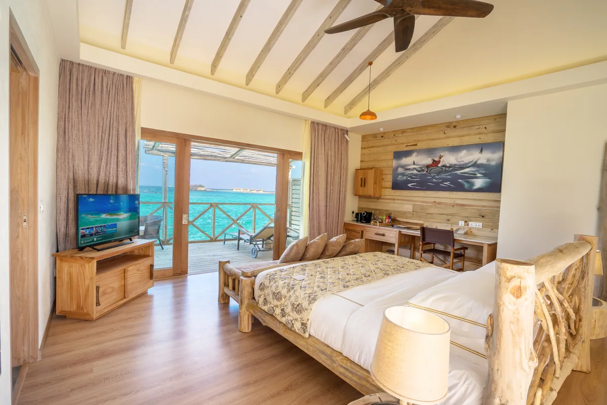 You & Me Maldives by Cocoon Collection, villa beach, Raa Atoll, Maldives, Lagoon Villa bedroom interior, handcrafted driftwood-style natural-wood bed frame (rustic-chic signature), exposed pale-timber rafters above, a flying-koi-fish painting set into the wood-paneled feature wall, retractable doors opening to the deck and ocean view, study desk and TV console along the perimeter.