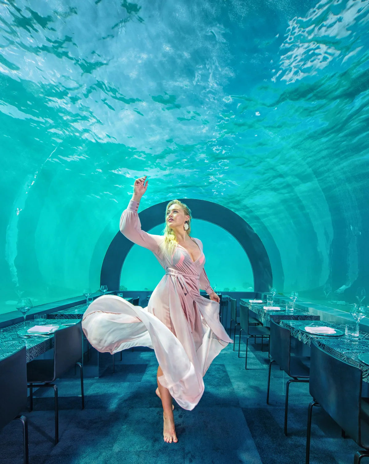 You & Me Maldives by Cocoon Collection, dining, Raa Atoll, Maldives, H2O by Andrea Berton aisle perspective: curved acrylic-tube architecture below the lagoon waterline, glass ceiling refracting turquoise light overhead with caustic patterns, model in pale-pink flowing dress walking down the central aisle between two rows of set tables, arched entryway at the tunnel end. The Michelin-tier underwater restaurant with the chef's Italian-influenced tasting menu.