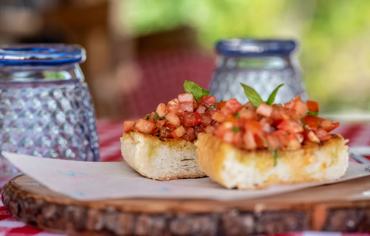 You & Me Maldives by Cocoon Collection, dining, Raa Atoll, Maldives, La Pasta Italian culinary close-up, two pieces of grilled bruschetta topped with diced tomato and basil chiffonade on a slatted wooden round board, cobalt-blue and pale-blue patterned drinking glasses behind, gingham-red tablecloth beneath, foliage-dappled garden light bokeh. The Cocoon Collection Italian regional kitchen as the broader-brand-DNA dining anchor.