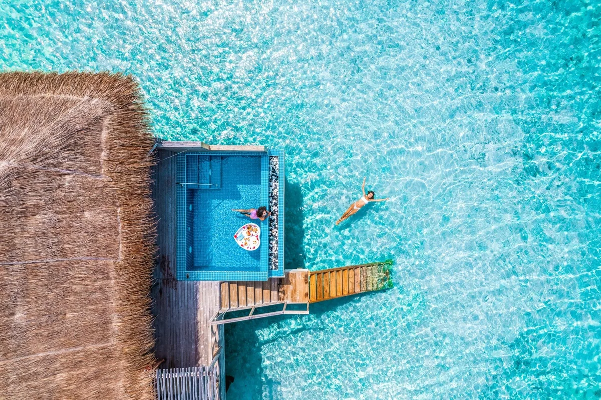 You & Me Maldives by Cocoon Collection, villa overwater, Raa Atoll, Maldives, Lagoon Suite with Pool top-down aerial, woman lounging in the small turquoise plunge pool inset into the deck adjoining the thatched villa, a second guest swimming in the surrounding lagoon, the deck access walkway extending to the right.