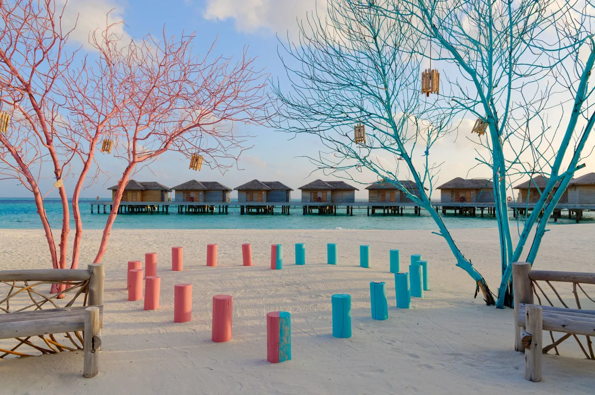 You & Me Maldives by Cocoon Collection, setting, Raa Atoll, Maldives, Painted-tree beach ceremony installation, pink-and-turquoise-painted decorative tree-branch sculptures at frame-left and frame-right, painted log stools in pink and teal-blue arranged in a circle on white sand around a central bench, the property's overwater villa row visible across the lagoon in middle distance, dusky-pink sky transitioning to pale-blue lagoon. Rustic-chic interior design language with handcrafted wooden furnishings.